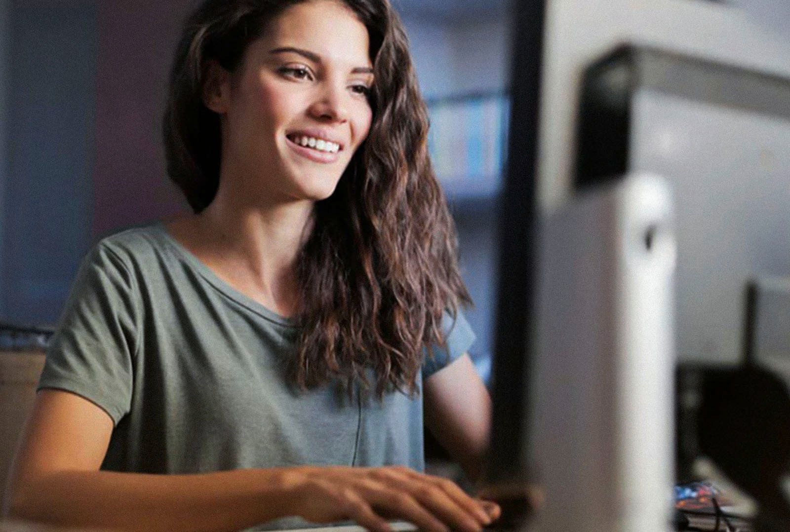 Junge Frau mit langen braunen Haaren lächelt und arbeitet an einem Desktop-Computer – symbolisiert moderne Weiterbildung und digitale Schulungen im Bereich Elektromobilität