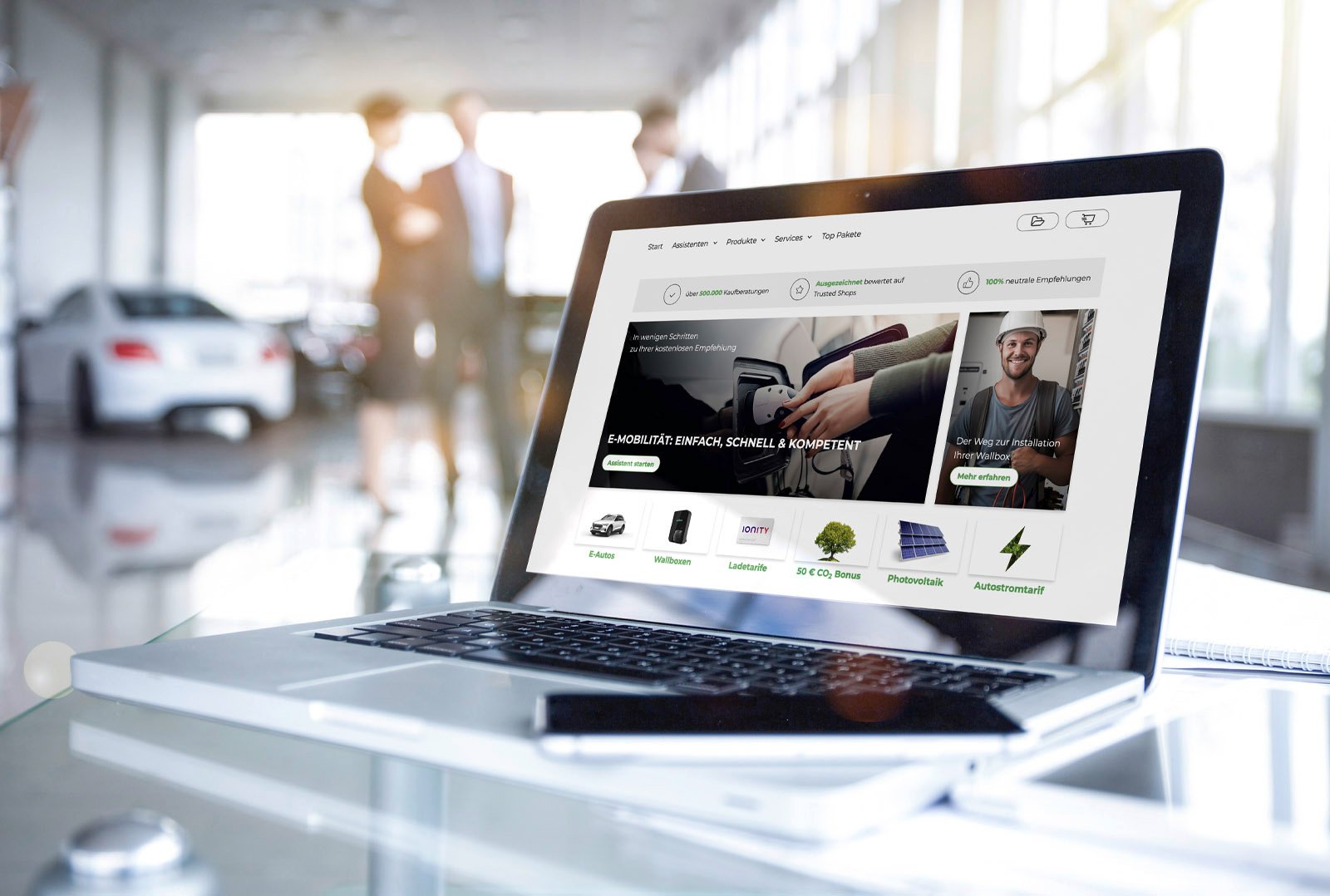 Laptop auf Glastisch mit geöffnetem e-mobilio E-Kaufberater, im Hintergrund Autohaus mit unscharfen Personen – Symbol für digitale Verkaufsberatung zur Elektromobilität im Privatkundenvertrieb